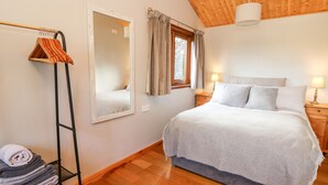 A bedroom with a bed, a mirror, a window with curtains, a wooden floor, and a nightstand with a lamp. - Y Caban Glas (Llandysul)