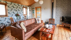 A cozy living room with a brown leather sofa, wooden coffee table, and a television. There is a patterned wallpaper with birds, a wooden ceiling, and a fireplace. - Y Caban Glas (Llandysul)