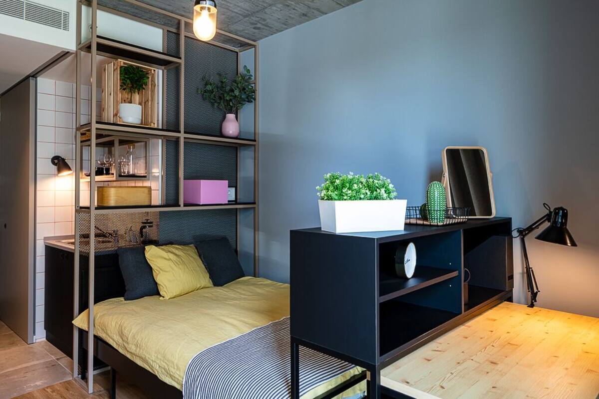 A compact bedroom with a bed, a desk, and shelves with plants and books.