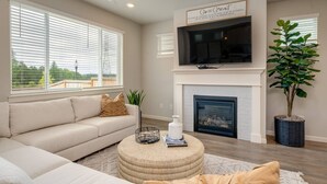 A living room with a beige sofa, a round ottoman, a fireplace, a large TV, and a potted plant. - Relaxing Getaway w/ Pool Access!

 (Camas)