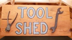 Een houten bord met de tekst 'TOOL SCHED' en twee gereedschappen die eronder hangen.