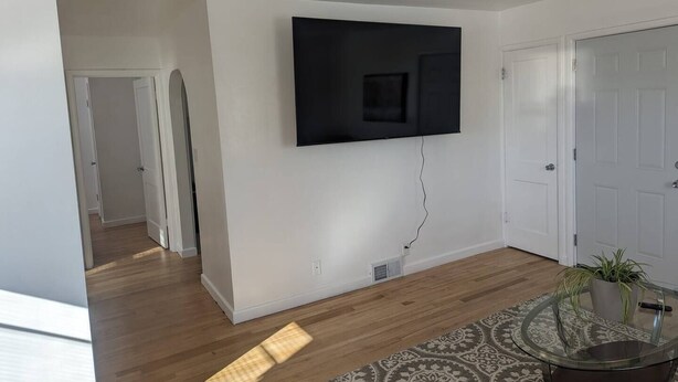 A modern living room with a flat-screen TV mounted on the wall, hardwood flooring, a patterned rug, and a glass table with a plant. - Nestled in the heart of Pierre, shopping & dining near. (Pierre)
