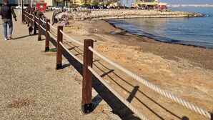 A coastal promenade with a stone walkway, palm trees, and a building with a red cross sign. - Caballito del Mar apartment in Punto Prima, Valencia (Punta Prima)