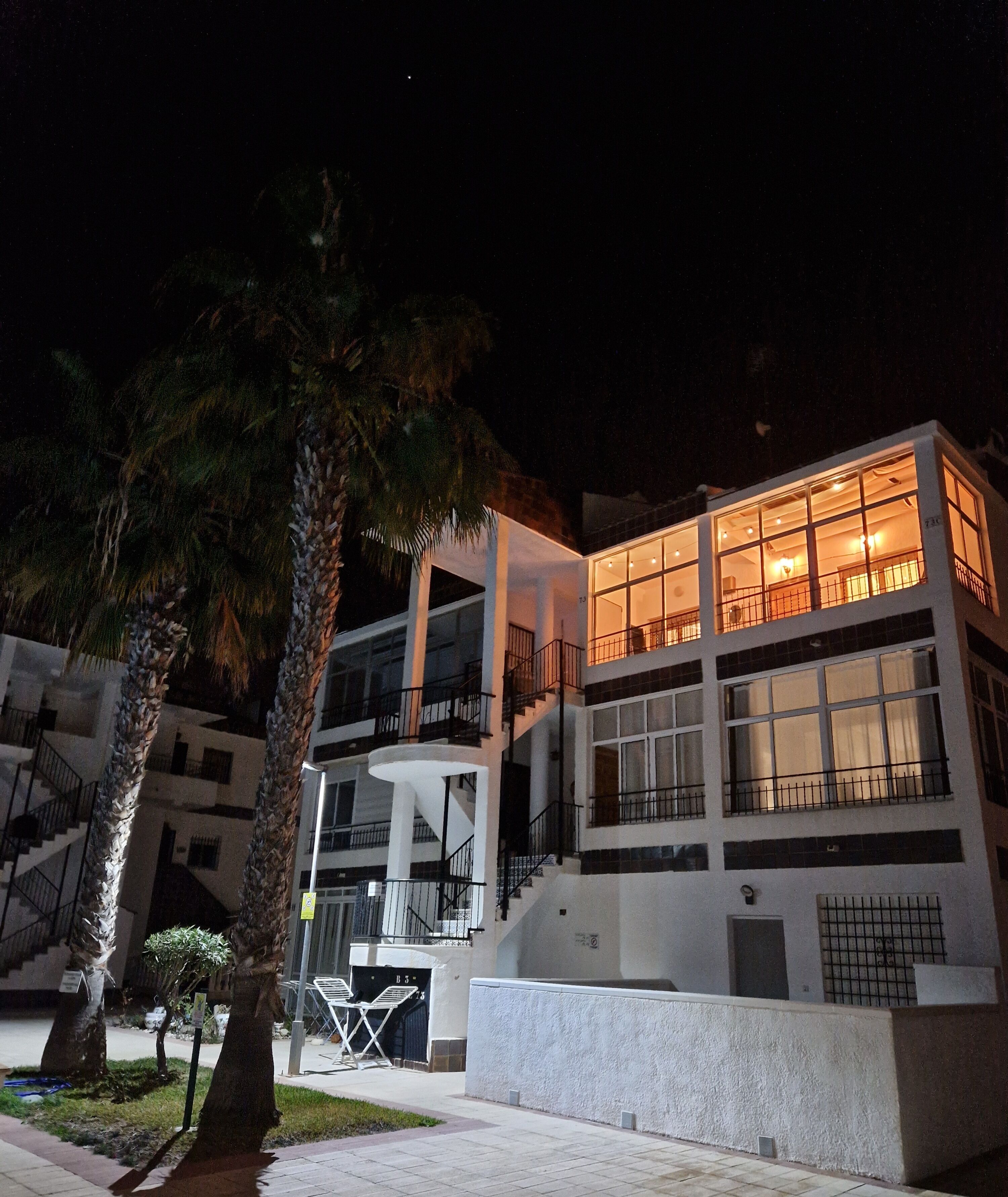 A multi-story building with balconies and large windows, surrounded by palm trees.
