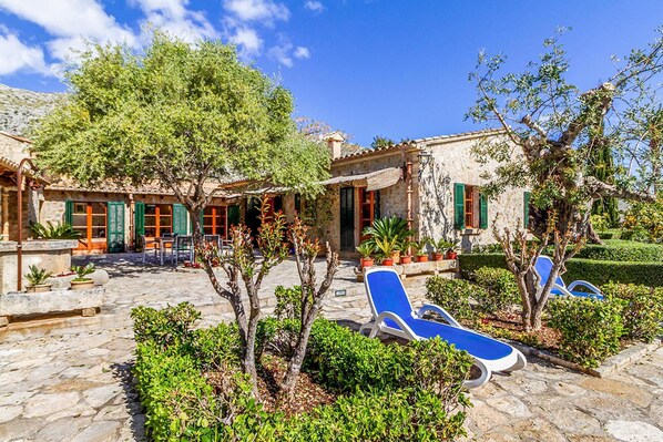 A stone courtyard with lounge chairs, a pergola, and a building with green shutters.