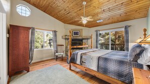 Un dormitorio con techo de madera, cama con colcha acolchada, chimenea de piedra y vistas a árboles a través de amplias ventanas.