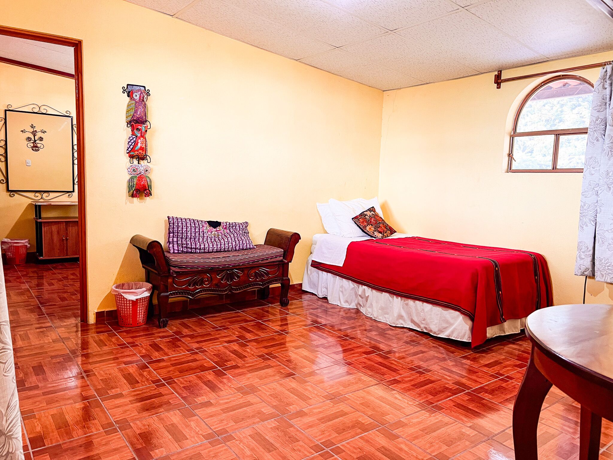 A room with a wooden floor, a bed with a red bedspread, a wooden chair, a small table, and a window with a curtain.