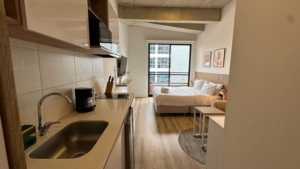 Eine moderne Küche mit Spüle, Kaffeemaschine und einem Messerblock. Ein Bett mit Kopfteil und Kissen, und ein Fenster mit Blick auf Gebäude.