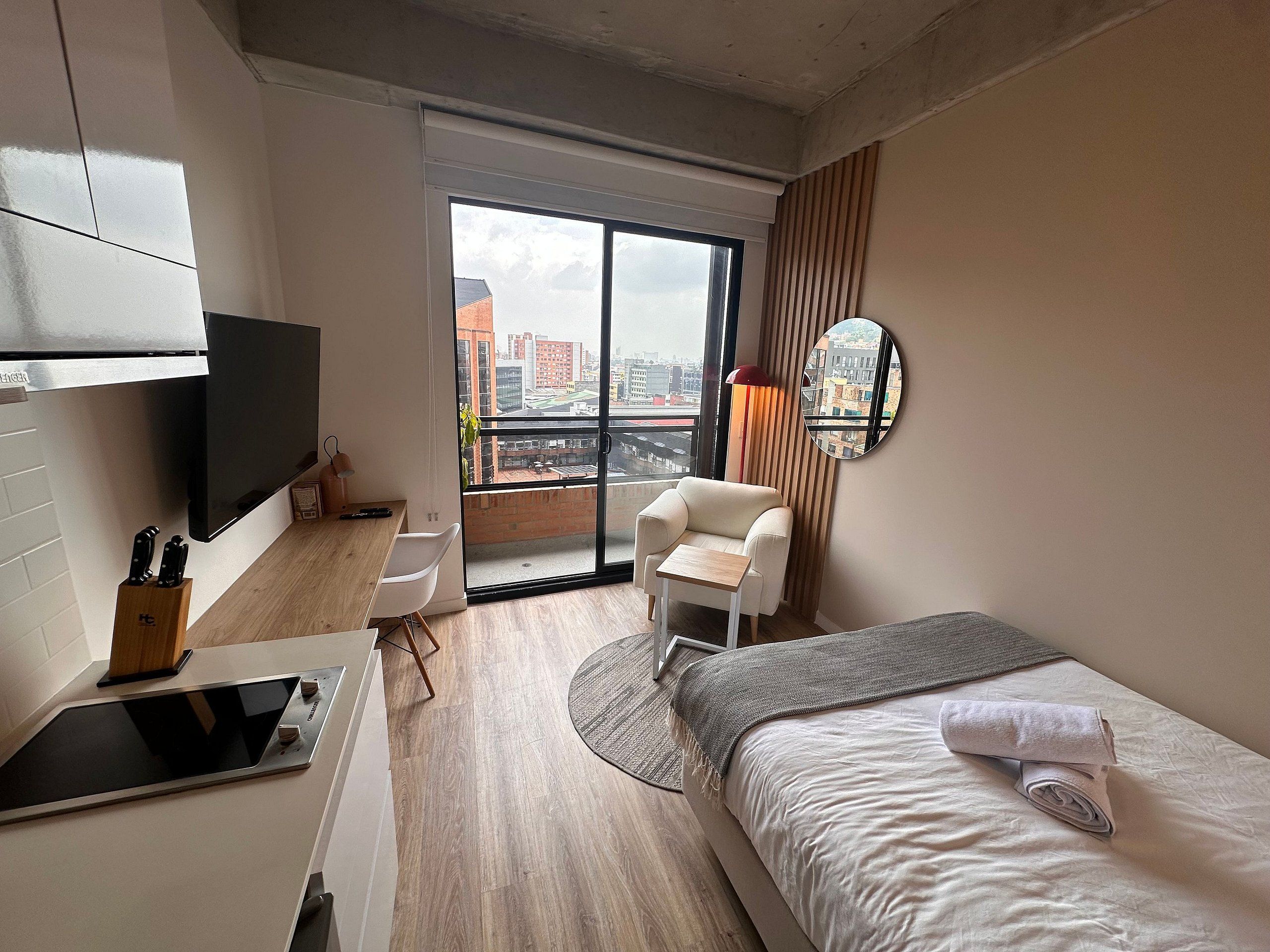 A modern hotel room with a bed, a desk with a flat-screen TV, a chair, a small table, and a large window with a city view.