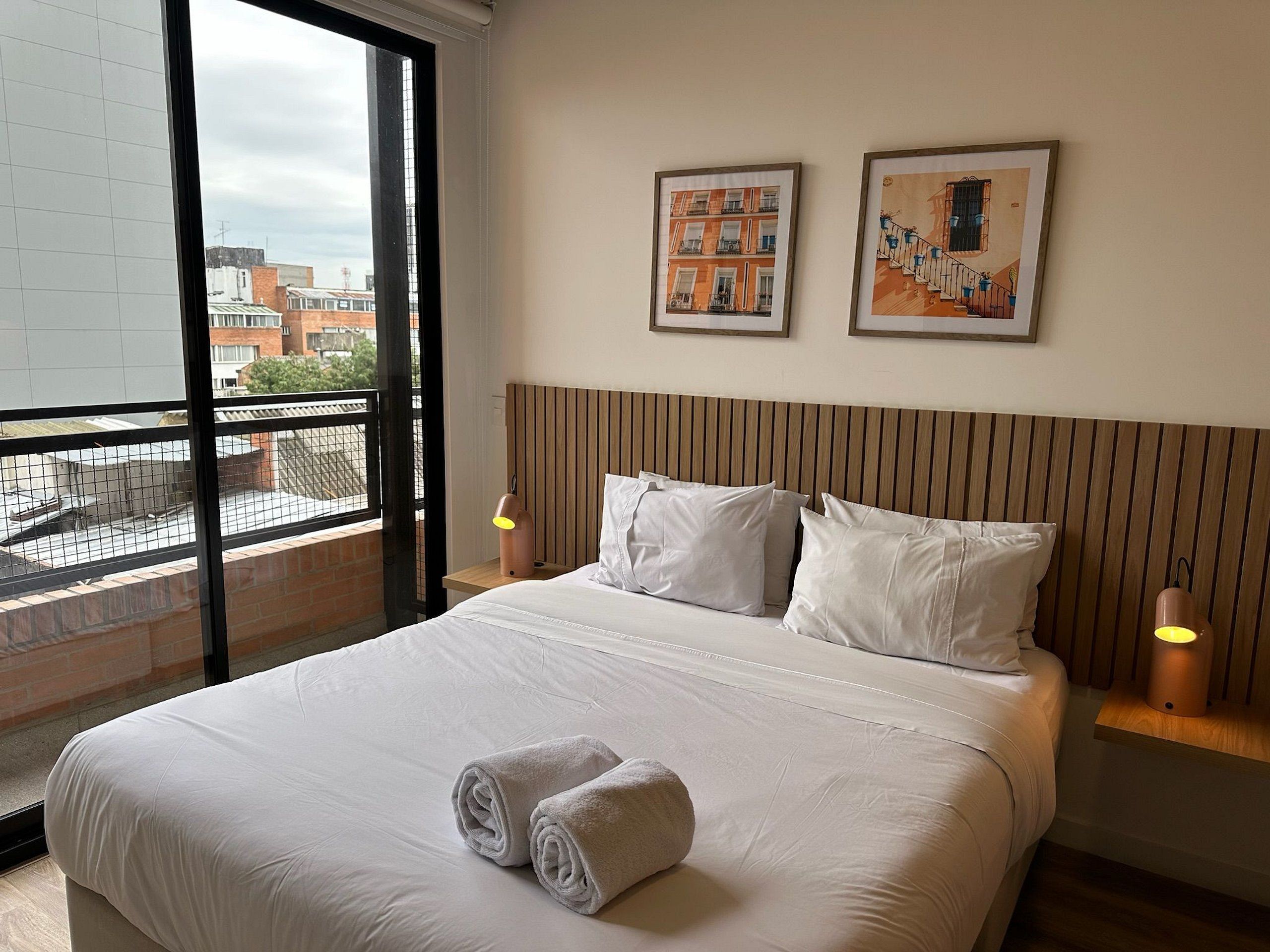Une chambre d’hôtel avec un grand lit, deux oreillers, une tête de lit, une fenêtre et deux tableaux accrochés au mur.