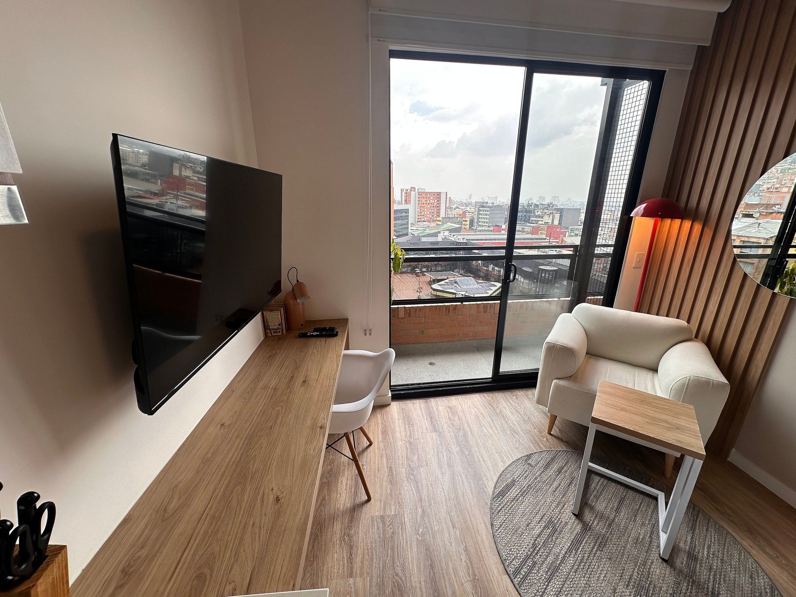 A modern living room with a flat-screen TV, a sofa, a small table, and a large window with a city view.