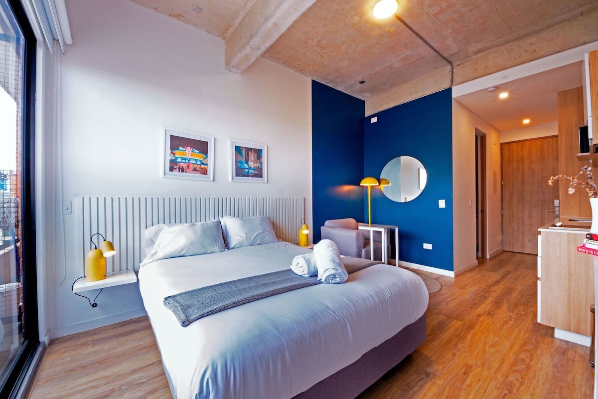 Une chambre d’hôtel moderne avec un grand lit, une lampe de chevet, une petite table, une chaise, un miroir et des œuvres d’art accrochées au mur.