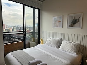 A hotel room with a large window, a bed with white linens, two framed artworks on the wall, and a potted plant on the windowsill.