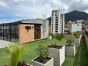 A rooftop terrace with artificial grass, concrete planters with plants, and a modern building with large windows and a brick facade.