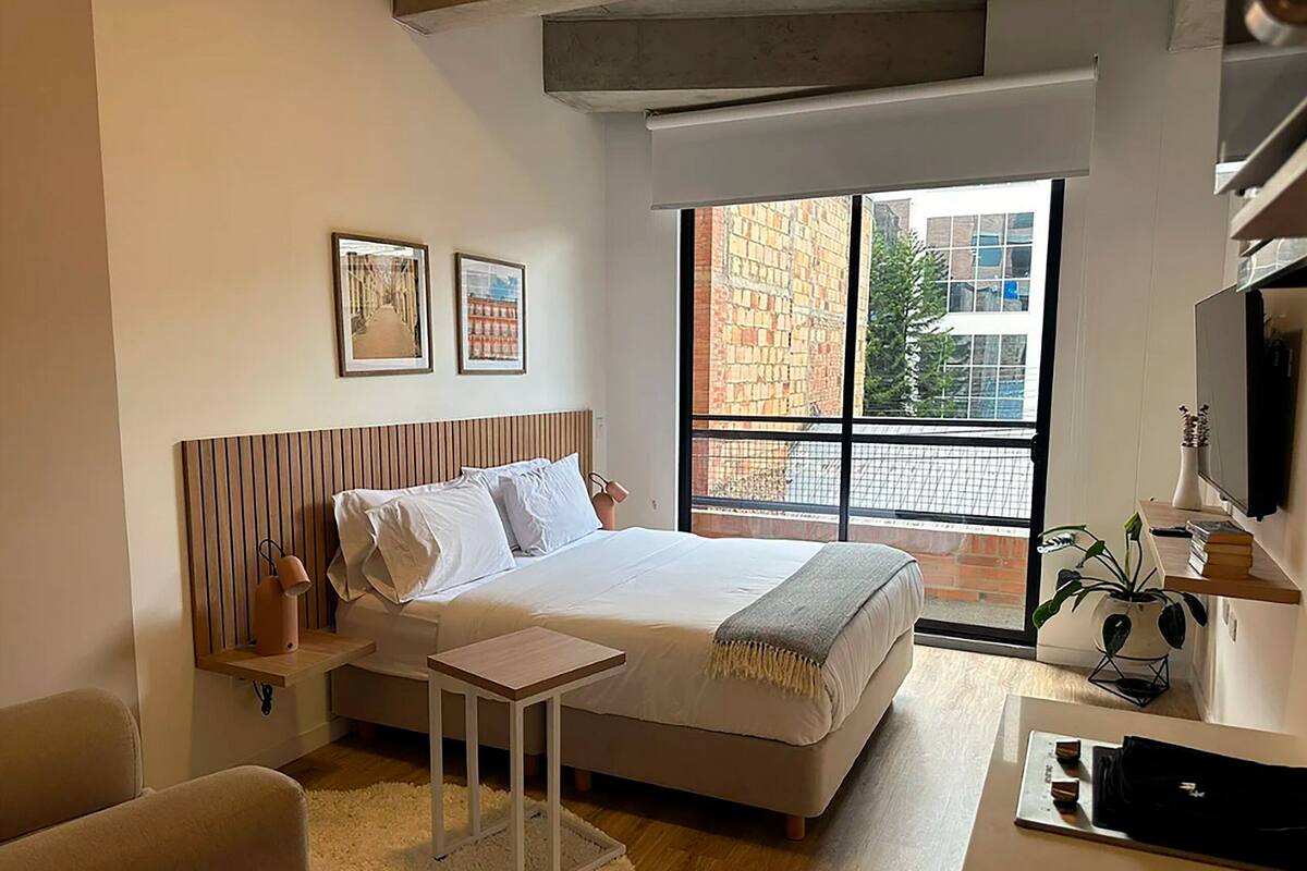 Une chambre moderne comprenant un lit, des tables de chevet, une petite table, une chaise, un bureau, une plante en pot et une fenêtre donnant sur des immeubles.