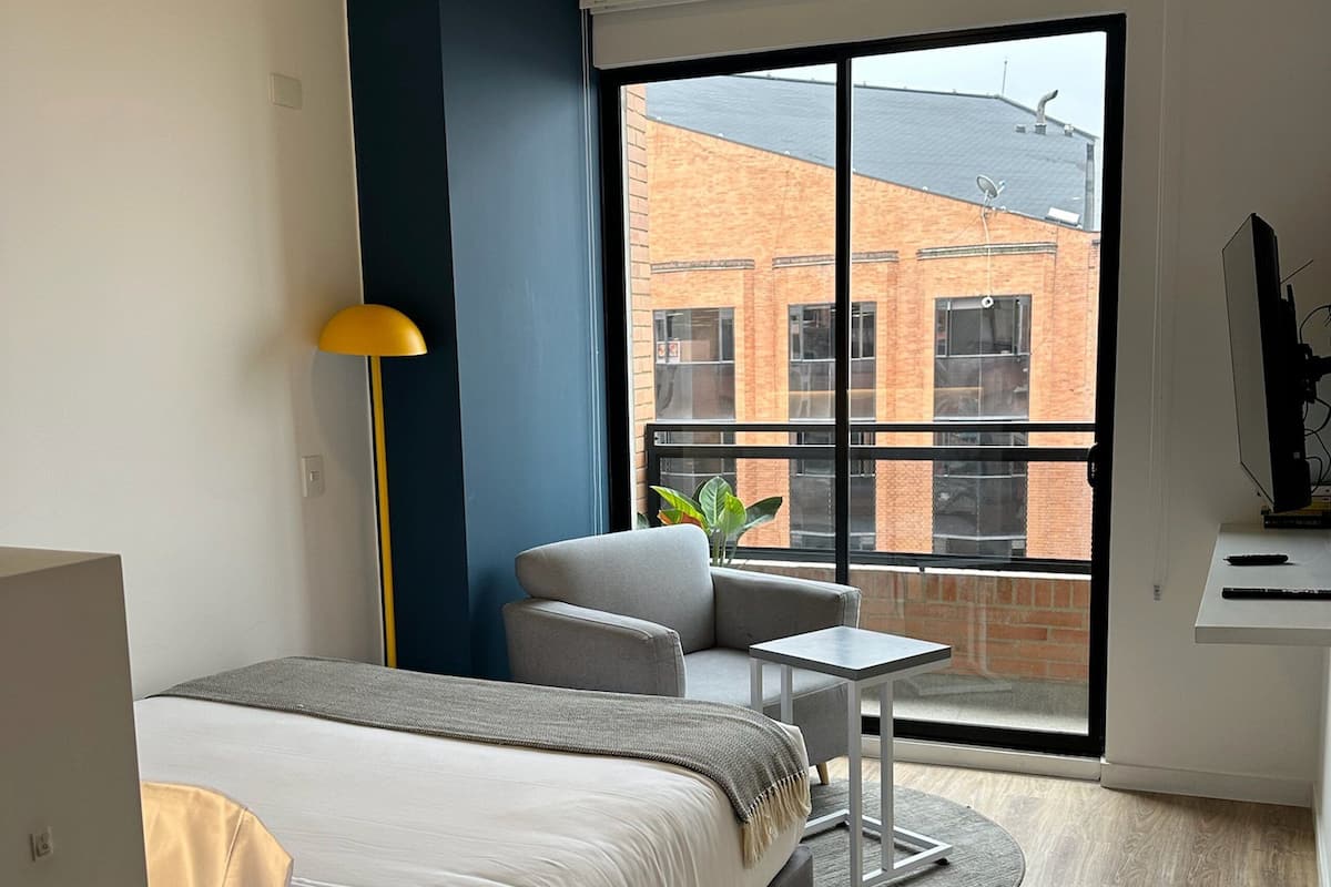 Une chambre d’hôtel moderne équipée d’un lit, d’une petite table, d’une chaise, d’un bureau, d’une lampe et d’une grande fenêtre donnant sur un immeuble en briques.