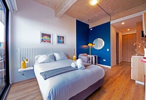 Une chambre d’hôtel moderne avec un grand lit, une lampe de chevet, une petite table, une chaise, un miroir et des œuvres d’art accrochées au mur.