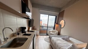 Ein kompaktes Hotelzimmer mit Küchenzeile, einem Bett, einer Sitzecke und einem großen Fenster mit Blick auf die Stadt.
