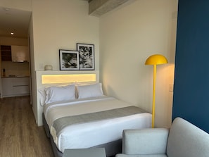 A hotel room with a bed, bedside table, lamp, and a kitchenette in the background.