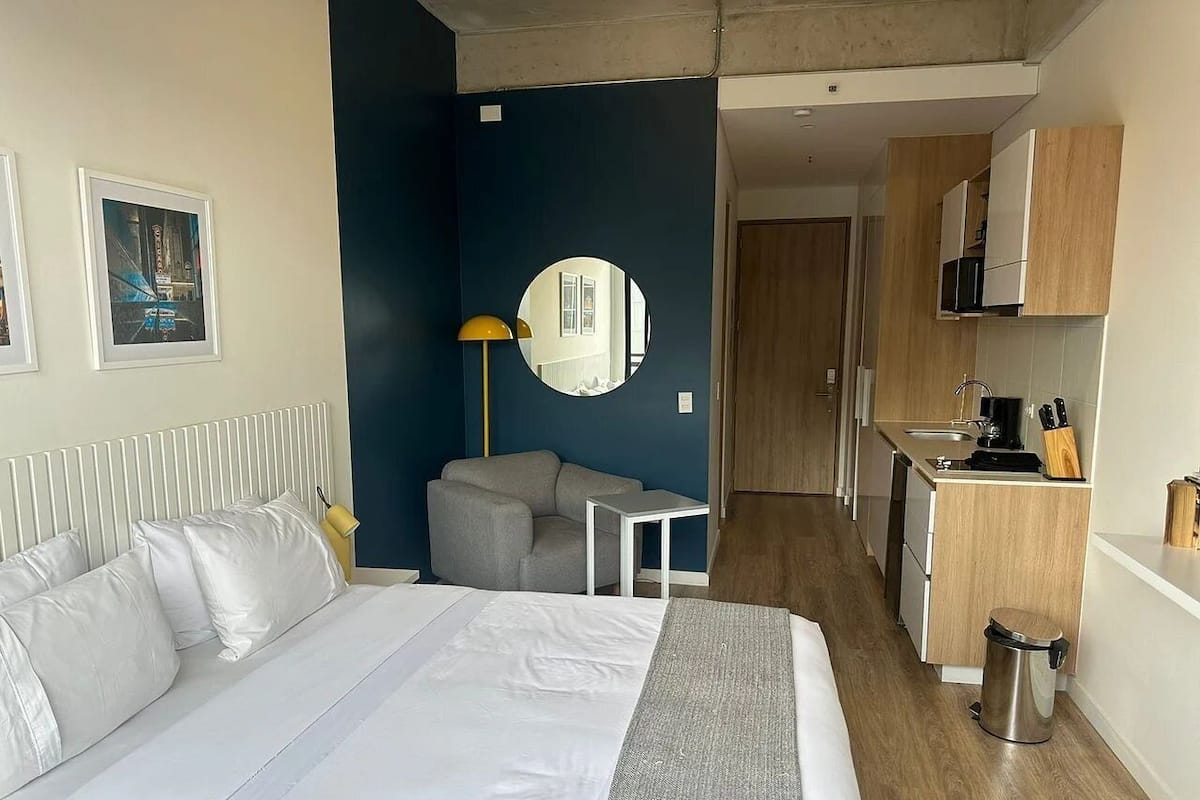 Une chambre d’hôtel comprenant un lit, un petit bureau, une chaise, un miroir rond, une kitchenette et une porte donnant sur une autre pièce.