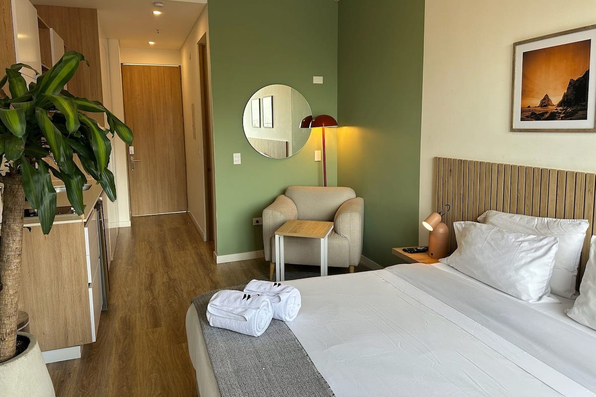 Une chambre d’hôtel avec un lit, une chaise, une petite table, un miroir, une lampe et une plante en pot.