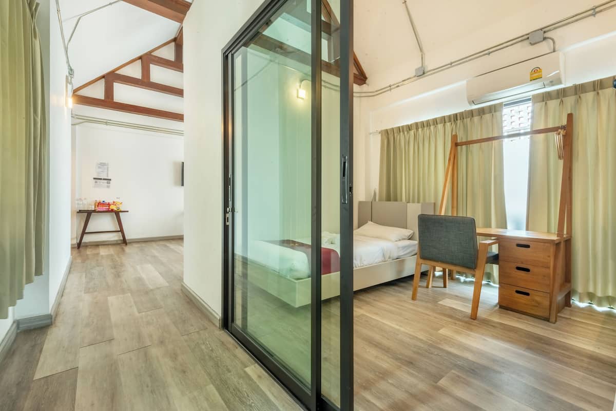 Habitació moderna amb llit, escriptori i cadira, terres de fusta i una porta corredissa de vidre que dona a una altra habitació.