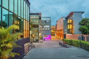 Un bâtiment moderne doté de grandes fenêtres, d’une entrée illuminée par des néons et d’un espace extérieur bien entretenu, agrémenté de plantes.