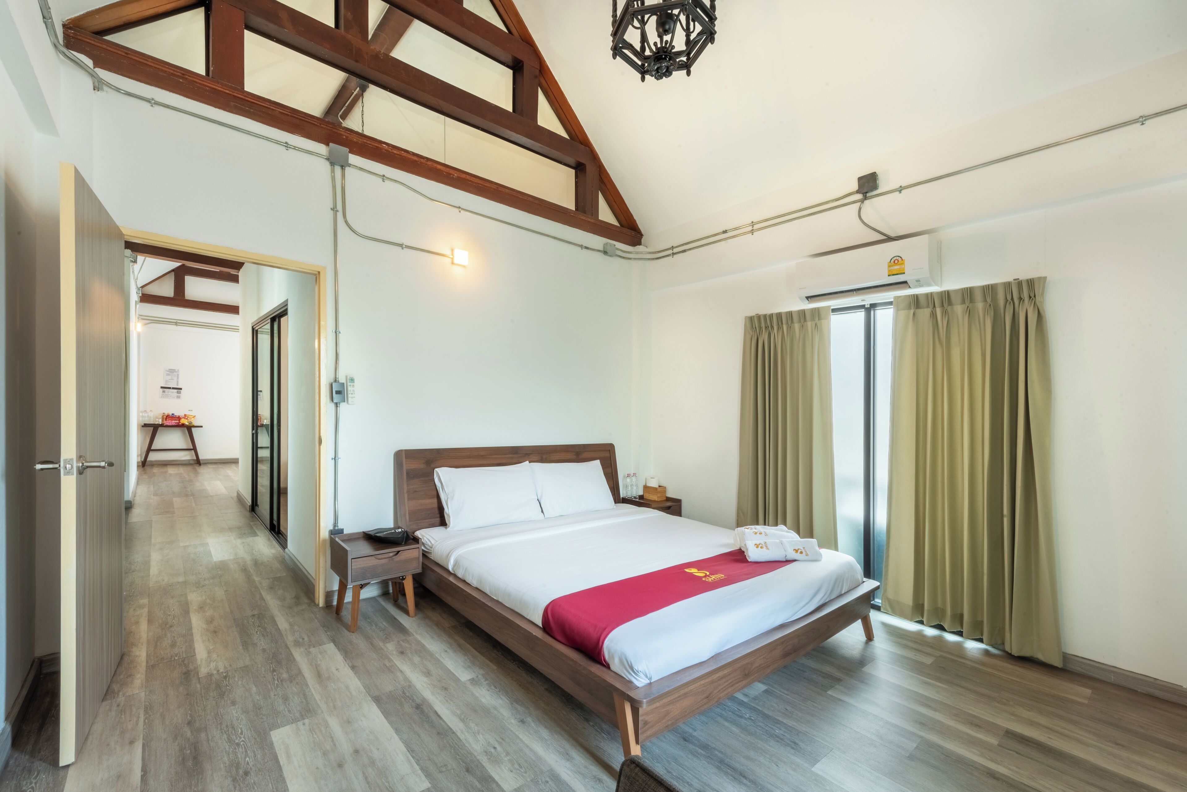 A hotel room with a wooden bed, a bedside table, a nightstand, a window with curtains, and a hallway leading to another room.