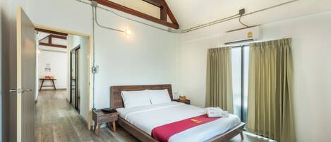 A hotel room with a wooden bed, a bedside table, a nightstand, a window with curtains, and a hallway leading to another room.