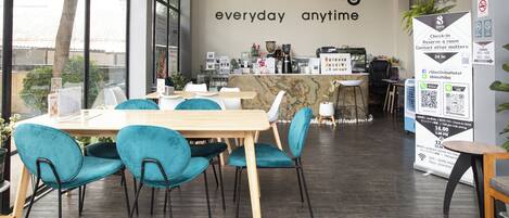 A modern cafe interior with a large window, teal chairs, a wooden table, and a wall sign reading "Caffeine King everyday anytime".