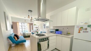 A modern kitchen with white cabinets, a built-in stove, and a refrigerator. There is a blue sofa in the living area and a window with curtains. - Exclusive Apartment Just Steps from San Isidro (Lince)