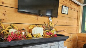 Ein Kamin mit Steinverkleidung, darüber ein Flachbildfernseher und ein hölzerner Sims, verziert mit Herbstlaub und Blumen.