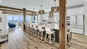 A modern kitchen with white cabinetry, a central island with bar stools, and a view of the ocean. - The Turtle's Palace- Oceanfront - Pool - Hot Tub - Dog Friendly (Surf City)