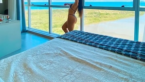 A bedroom with a large window, a bed with a patterned bedspread, and a view of the beach.