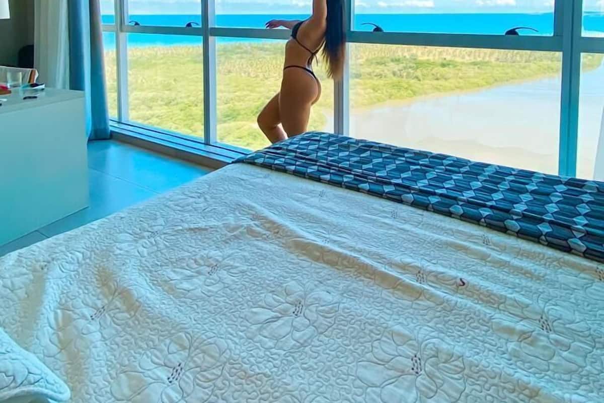 A bedroom with a large window, a bed with a patterned bedspread, and a view of the beach.