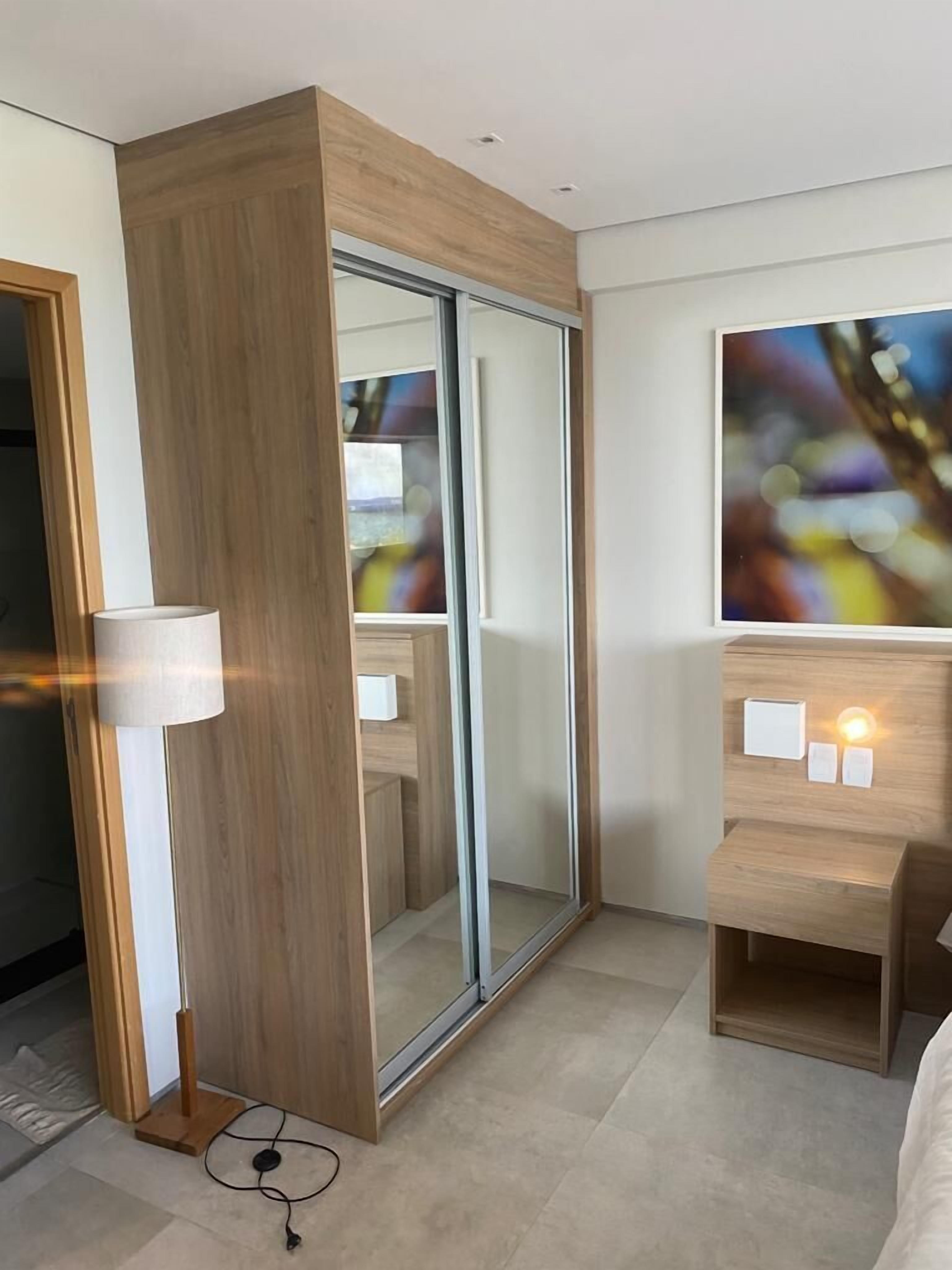Een moderne hotelkamer met een houten garderobe, een glazen deur, een nachtkastje met een lamp, en een schilderij aan de muur.