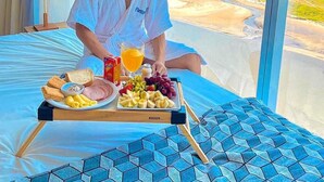 A person sitting on a bed with a tray of breakfast items, including toast, ham, and fruit, in a room with a large window overlooking a landscape.