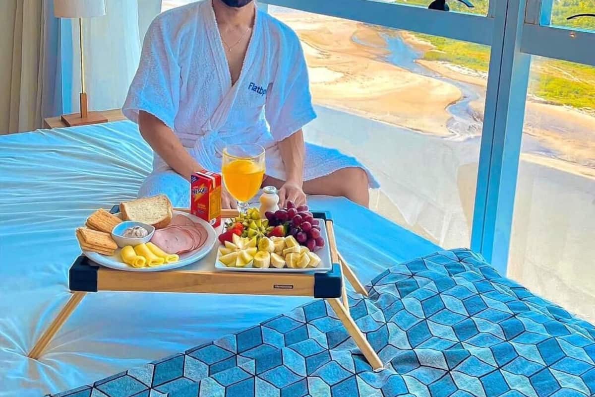 A person sitting on a bed with a tray of breakfast items, including toast, ham, and fruit, in a room with a large window overlooking a landscape.