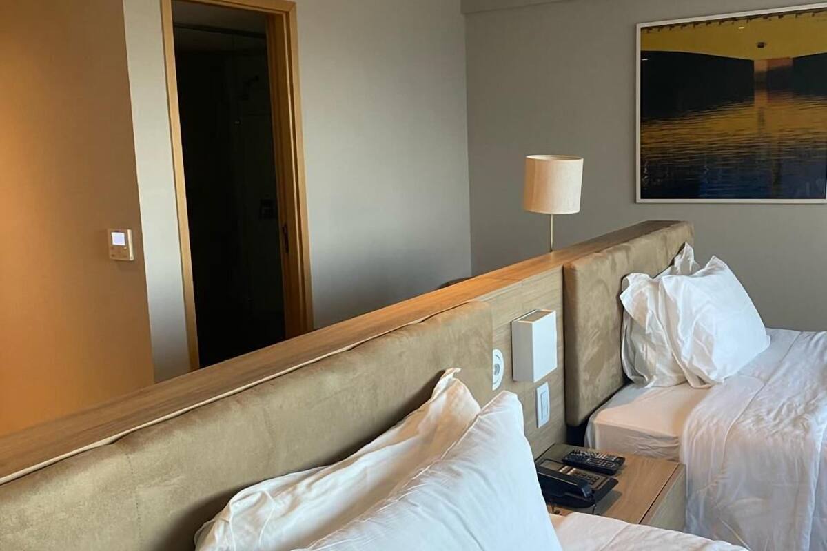 A hotel room with two beds, a bedside table with a lamp, a painting on the wall, and a door leading to another room.