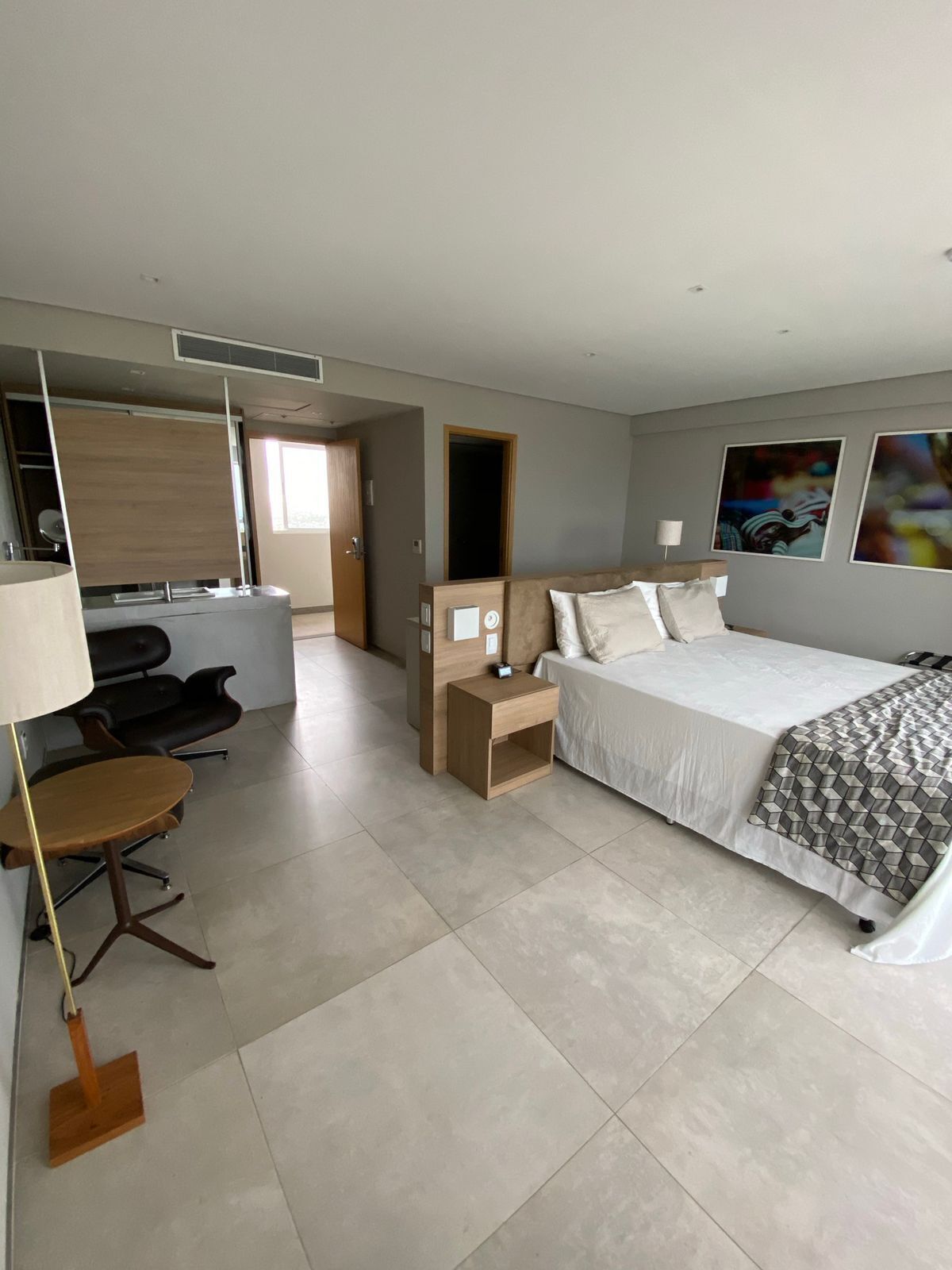 Quarto de hotel moderno com cama, mesa de cabeceira, escrivaninha, cadeira e uma janela grande.