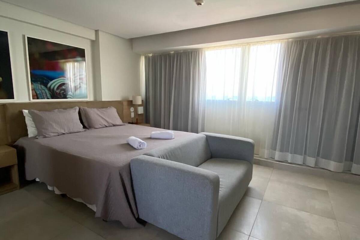 A modern hotel room with a large bed, a sofa, a nightstand, and a window with curtains.