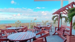An outdoor patio with wooden tables and benches, a covered seating area, and a view of the ocean.