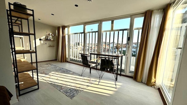 A spacious room with a large window, a wooden floor, a black metal shelf, a dining table with chairs, and a patterned rug. - Digital Nomad House (Istanbul)