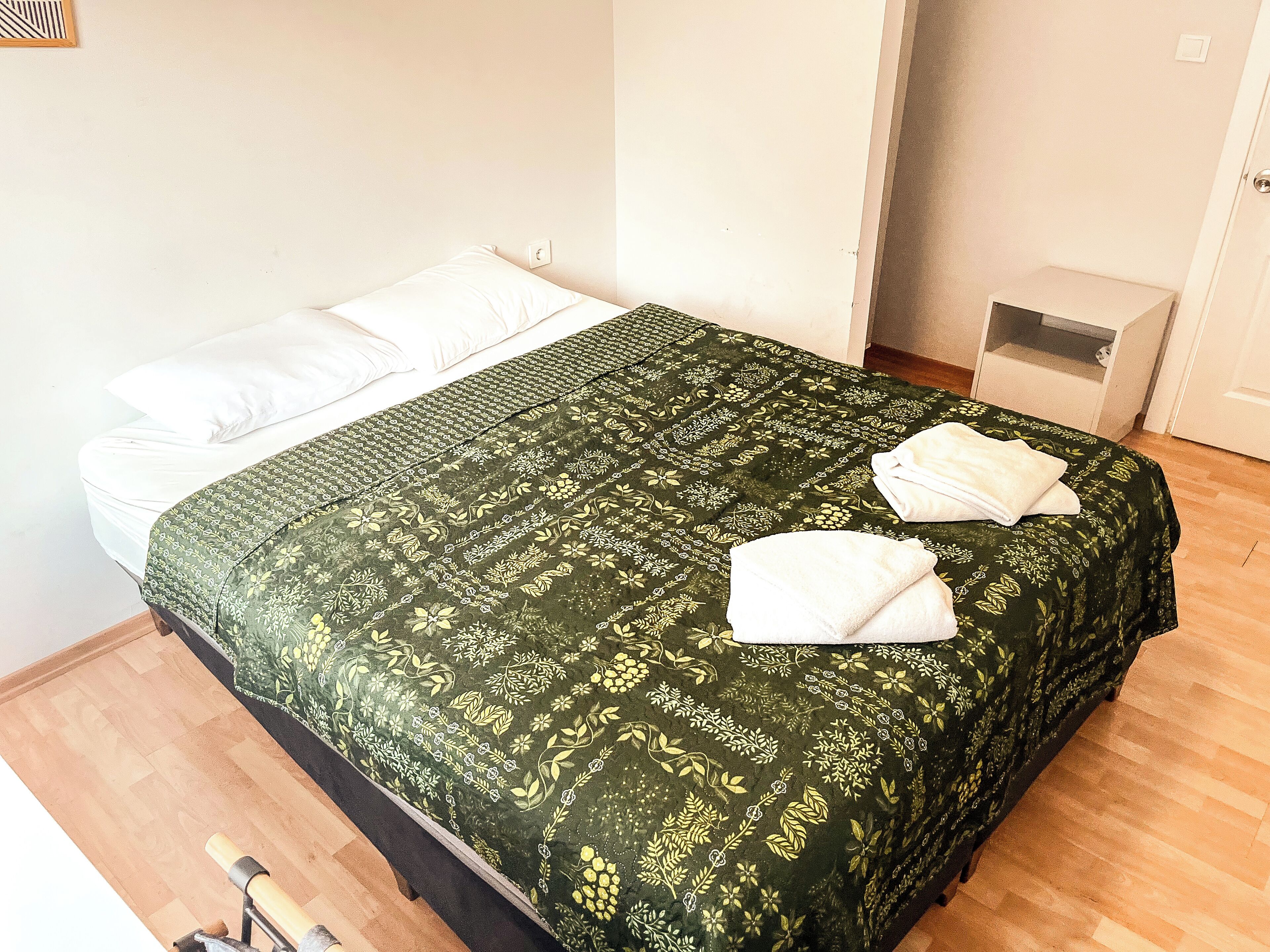 A bed with a green floral bedspread and white pillows, a wooden floor, and a small bedside table with a white lamp.