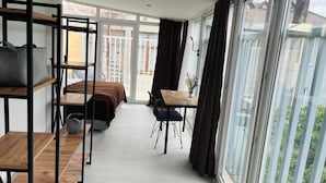 A modern, well-lit room with a large sliding glass door leading to a balcony, a bed with a wooden headboard, a small table with a chair, and a sofa with a patterned cushion. - Digital Nomad House (Istanbul)