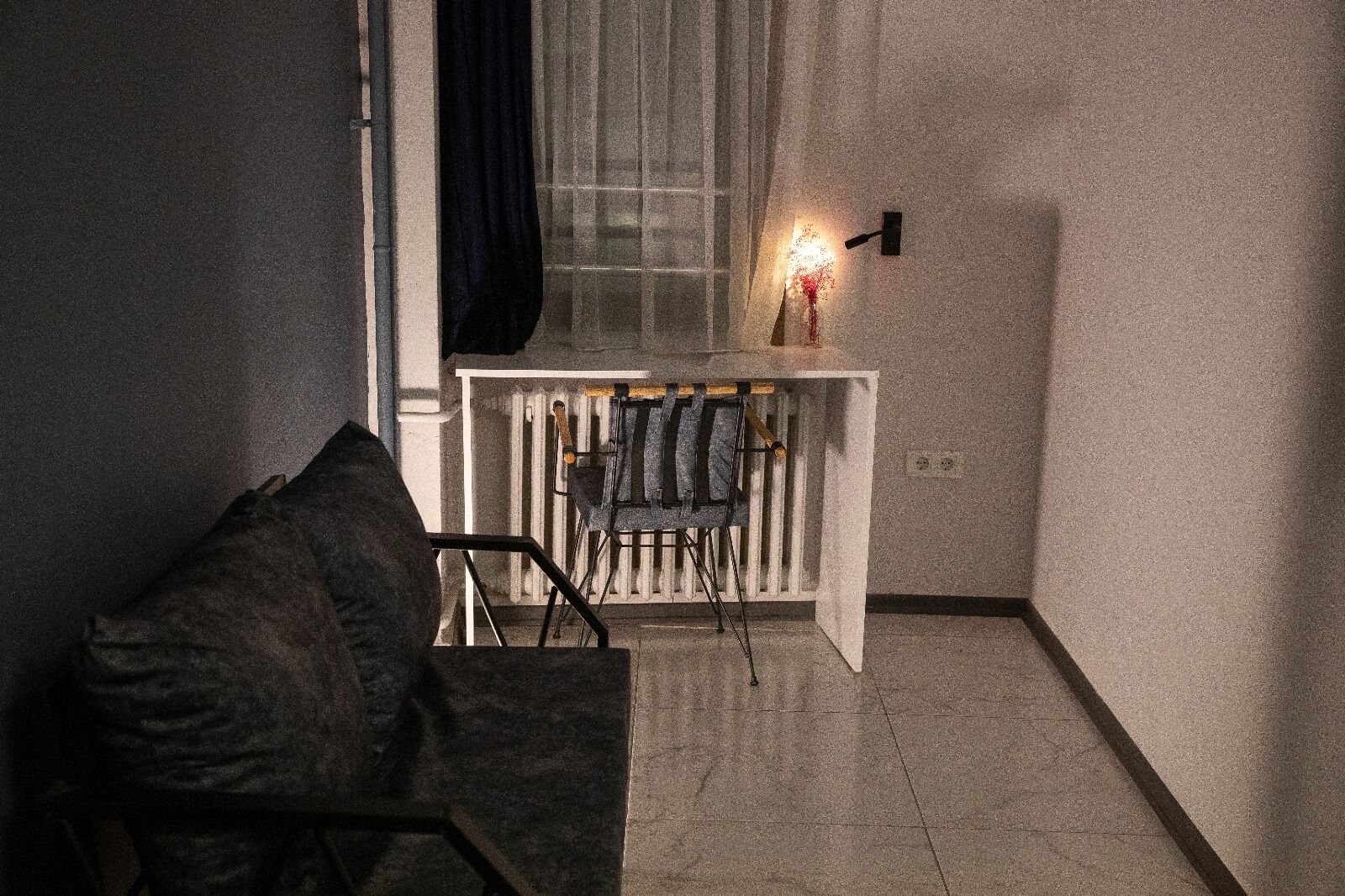 A dimly lit room with a white radiator, a black chair, and a small table with a wall-mounted light.