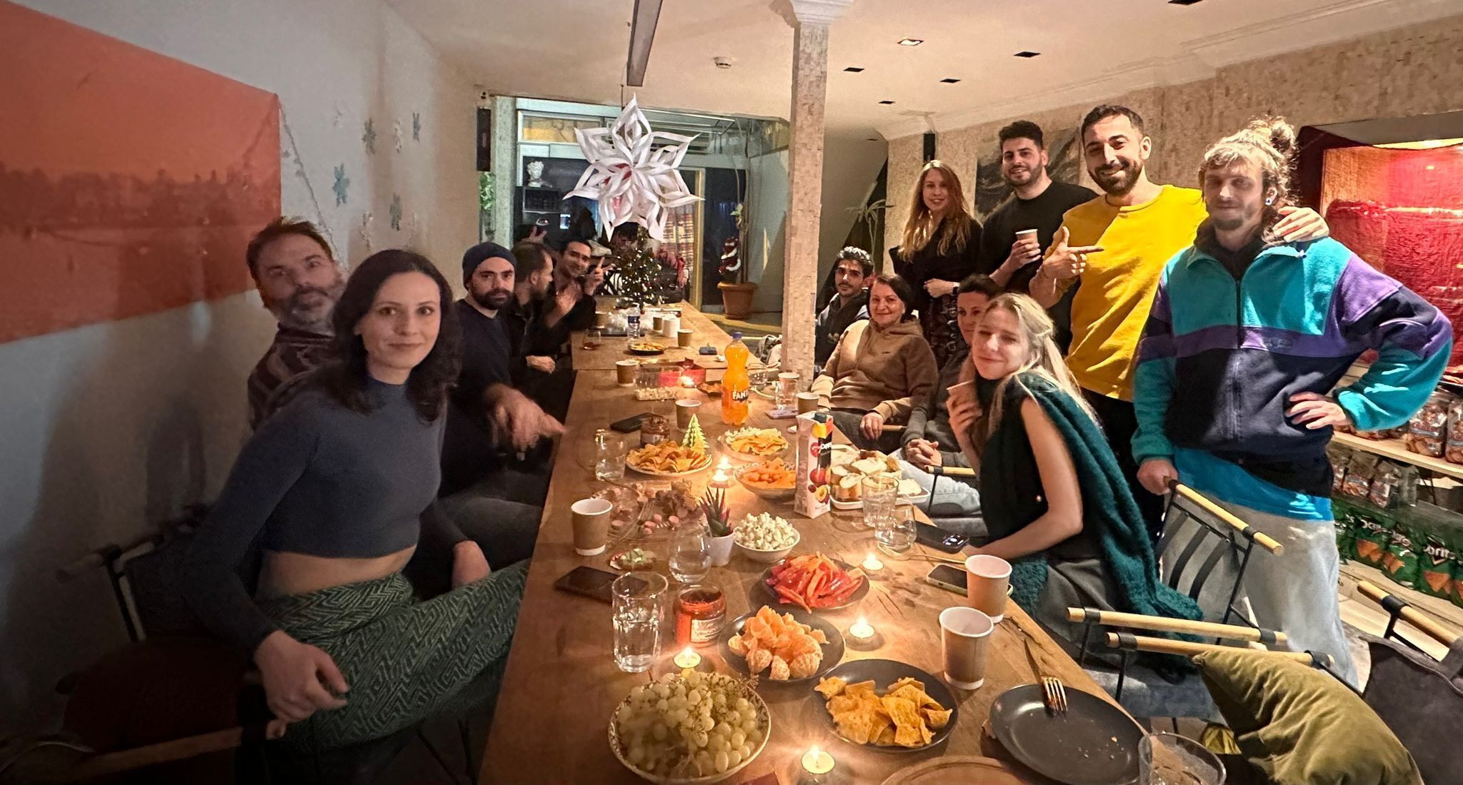 A group of people gathered around a long table with various food items and drinks, in a room with a large window and a decorative white star hanging from the ceiling.