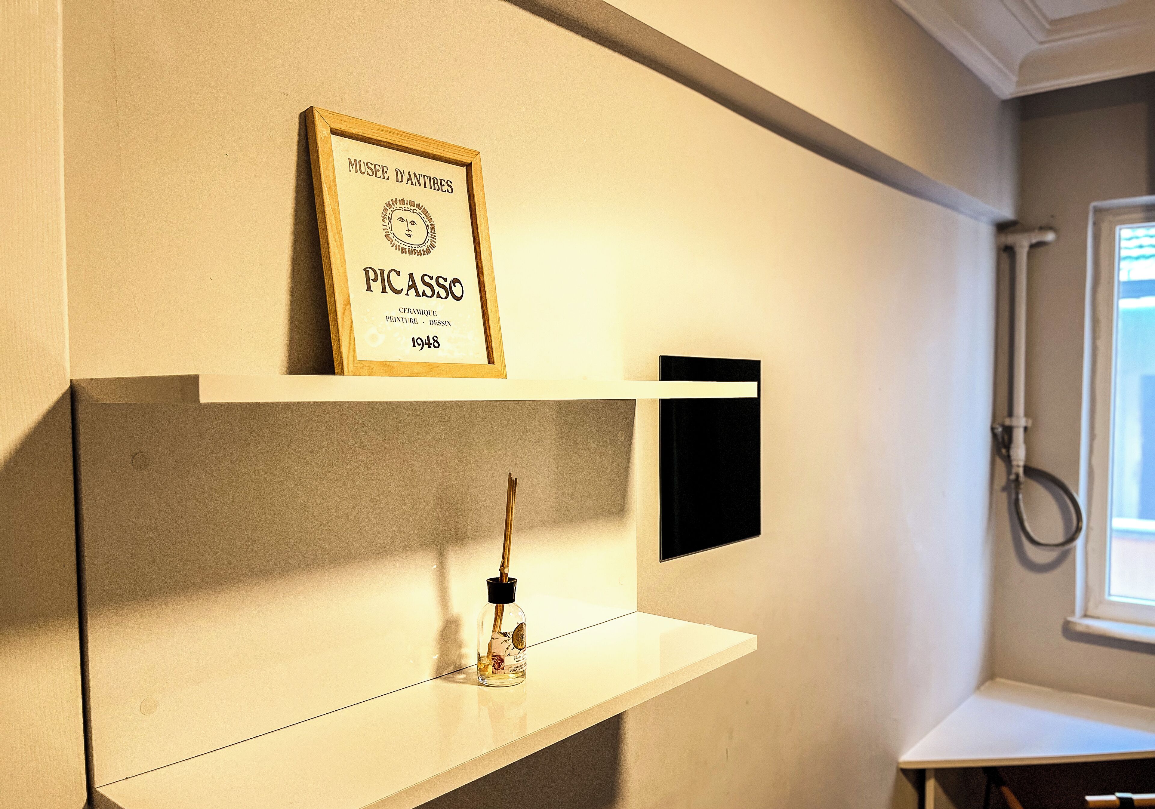 A framed Picasso artwork on a wall, a small shelf with a bottle, and a black object on a white shelf.