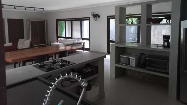 A modern kitchen with a central island, built-in appliances, and a dining area with a sofa and table. - Casa em Maresias em Meio a Mata (Praia de Maresias São Sebastião)
