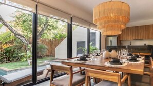 Un espace de salle à manger moderne, avec une grande table dressée pour six personnes, entourée de chaises en bois dotées d’assises rembourrées. Un grand luminaire suspendu au design original est placé au-dessus de la table.